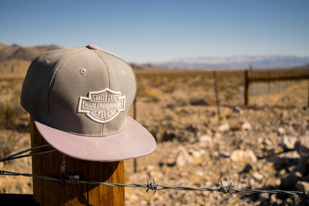 A Harley Davidson cap sits on a fence post in an arid desert landscape, under a bright blue sky.